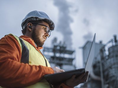 Ingeniero con casco de seguridad y chaleco reflectante trabajando en una laptop frente a una planta industrial, simbolizando monitoreo y análisis en tiempo real en entornos críticos.